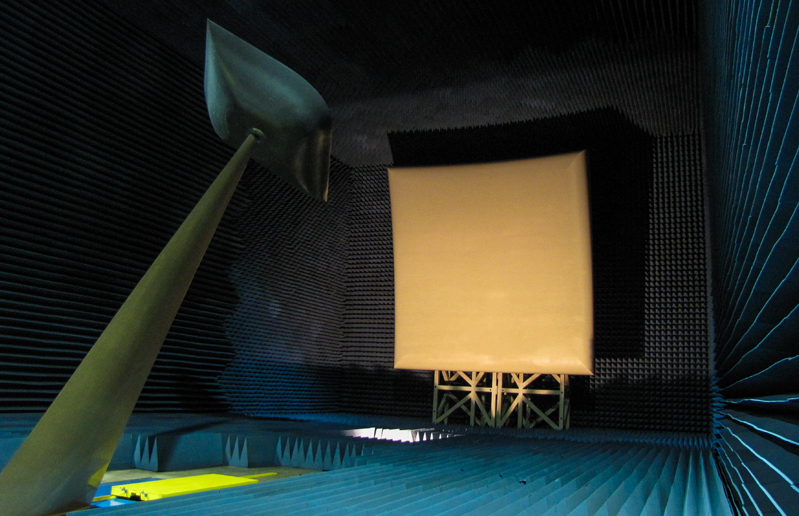 A photo of a Compact Antenna Test Range (CATR) facility. The image shows a shielded room with an anechoic chamber, a robotic positioning system, and various measurement equipment, including spectrum analyzers and network analyzers. The control room can be seen through the glass window, where engineers and technicians can monitor the test results. CATR is used for testing and evaluating the performance of small and medium-sized antennas.